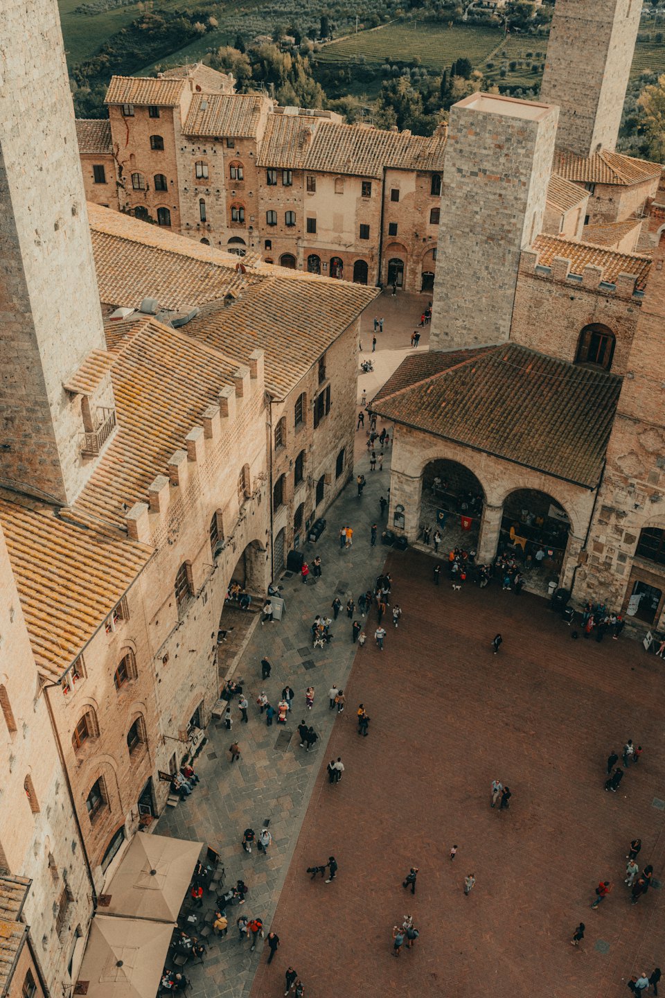Piazza storica con torri, caffè e colline verdi in lontananza.